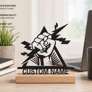 Peut inclure: Une enseigne en acrylique noir découpée au laser représentant un poing fermé entouré d'éclairs à l'intérieur d'un triangle. L'enseigne porte le texte CUSTOM NAME et est fixée sur une base rectangulaire en bois massif de couleur claire.