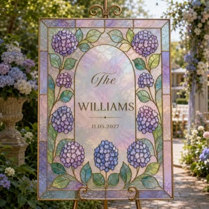 Può includere: Un cartello rettangolare in vetro colorato con finitura iridescente, caratterizzato da un bordo di fiori di ortensia viola e blu con foglie verdi. Il centro mostra il testo "The WILLIAMS" e la data "11.05.2027" in un carattere serif dorato.