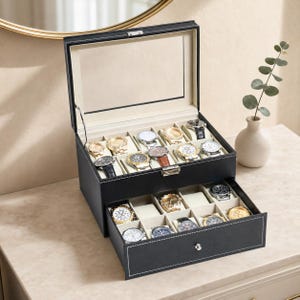 May include: A black watch box with a glass lid and a pull-out drawer, featuring white contrast stitching. The interior is lined with beige fabric and contains multiple compartments, each holding a wristwatch with various metal and leather bands.