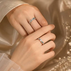 May include: Two silver rings featuring a delicate leaf motif with green and blue enamel detailing. One ring is a simple band, while the other is a bypass-style ring centered with a single, clear, round-cut gemstone set in a silver prong setting.