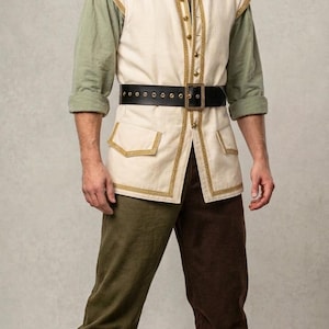 May include: A cream-colored sleeveless vest with gold trim, worn over a sage green long-sleeved shirt. The outfit includes a black leather belt with a gold buckle, two-toned trousers in olive and dark brown, and brown leather mid-calf boots.