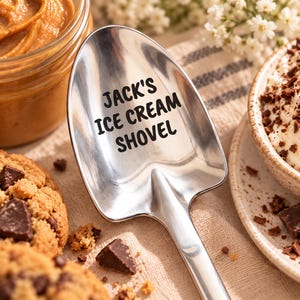 May include: A polished stainless steel shovel-shaped spoon featuring the black engraved text "JACK'S ICE CREAM SHOVEL" on the bowl. The utensil has a wide, flat-edged head and a smooth, reflective surface, designed for scooping frozen desserts.