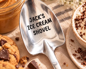 May include: A polished stainless steel shovel-shaped spoon featuring the black engraved text "JACK'S ICE CREAM SHOVEL" on the bowl. The utensil has a wide, flat-edged head and a smooth, reflective surface, designed for scooping frozen desserts.