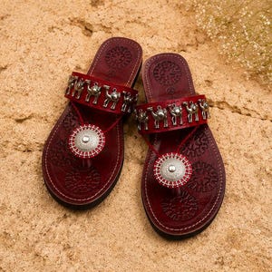May include: A pair of dark red leather sandals featuring a wide strap adorned with silver-toned camel charms. The footbed is embossed with circular patterns, and a central silver-toned circular medallion is attached to the toe post with red thread.