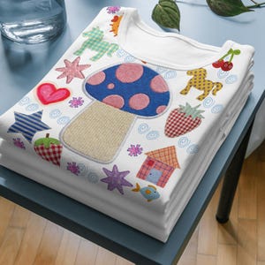 May include: A white t-shirt featuring a colorful pattern of a large blue mushroom with pink spots, red hearts, yellow and green horses, red strawberries, a small house, a blue fish, and various stars, all rendered in a playful, textured graphic style.