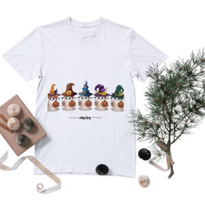 Puede incluir: Una camiseta blanca con un gráfico de cinco fantasmas que llevan sombreros de bruja coloridos y sostienen pequeñas calabazas naranjas. Debajo de los fantasmas, la palabra "mama" está impresa en cursiva negra junto a pequeños murciélagos.