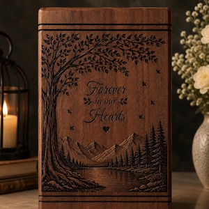 May include: A rectangular wooden cremation urn featuring a laser-engraved landscape scene. The design depicts a large tree with detailed leaves, a mountain range, a forest of pine trees, and a lake. The text Forever in our Hearts is centered.