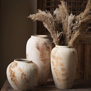 May include: Three ceramic vases with a textured, off-white finish and mottled brown, rust-coloured patches. The vases vary in height and shape, with the tallest containing a large arrangement of dried, feathery, beige pampas grass stalks.
