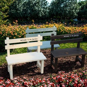 Può includere: Tre piccole panchine in legno di colore bianco, verde salvia e marrone scuro, ciascuna dotata di uno schienale con la scritta "Welcome to my Garden" stampata in un carattere semplice. Le panchine hanno una finitura usurata e poggiano sul pacciame.