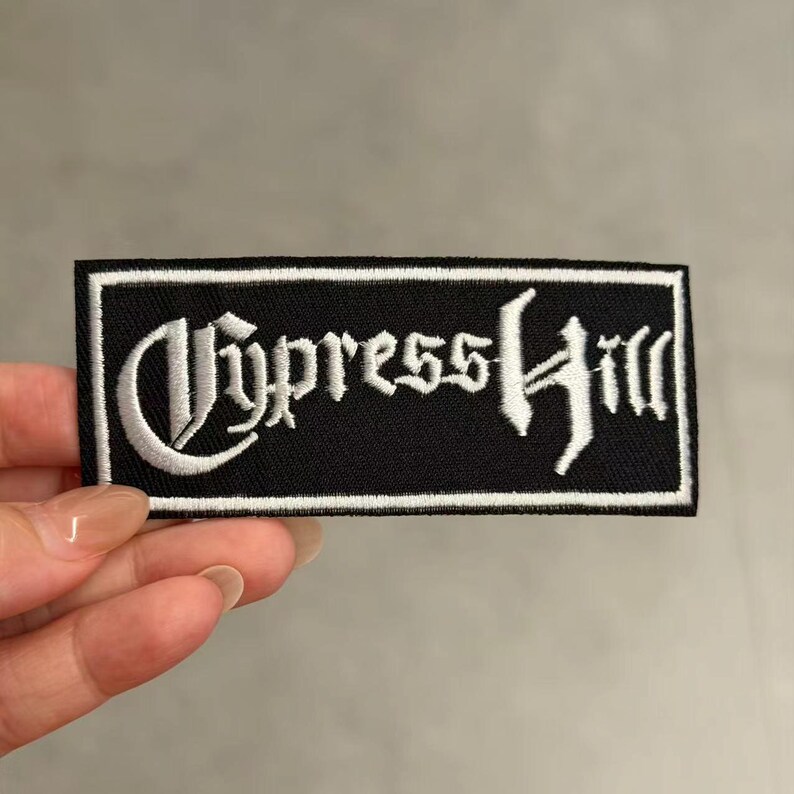 May include: A rectangular embroidered patch featuring a black background with a white border and the text Cypress Hill written in a stylised gothic font. The patch is designed for application to clothing or accessories with its clear white lettering.