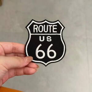 May include: A black and white embroidered patch in the shape of a shield, featuring the text ROUTE US 66 in white lettering. The design mimics the iconic highway sign with a bold white border and a solid black background for high contrast.