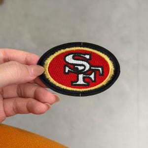 Puede incluir: Un parche bordado de forma ovalada con el logotipo de los San Francisco 49ers. El diseño incluye un centro rojo con las letras SF en blanco y negro, rodeado por un borde dorado y un borde exterior negro grueso con costuras detalladas.