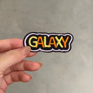May include: An embroidered patch with a black background and a wavy white border. The word GALAXY is stitched in the center using a gradient of yellow and orange thread. The patch has a rounded, irregular shape and a textured fabric appearance.