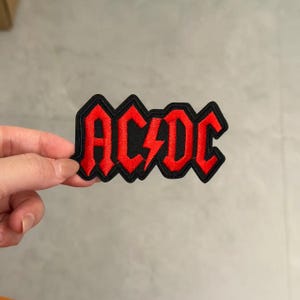 May include: An embroidered fabric patch featuring the band name AC/DC in bright red lettering with a central lightning bolt symbol. The text is outlined in a thick black border, creating a bold, high-contrast design suitable for application.