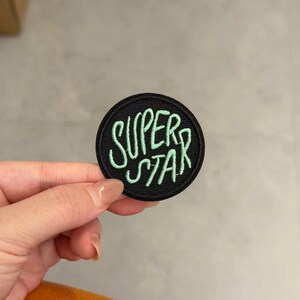 Pode incluir: Um emblema bordado circular com fundo preto e a palavra SUPERSTAR costurada em fio verde claro. O texto está disposto em duas linhas com uma fonte manuscrita estilizada, e o emblema apresenta uma borda preta costurada ao redor.