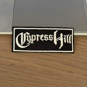 May include: A rectangular embroidered patch featuring a black background with a white border and the text Cypress Hill in a stylised gothic font. The patch is designed for application to fabric items and displays high-contrast white thread embroidery.