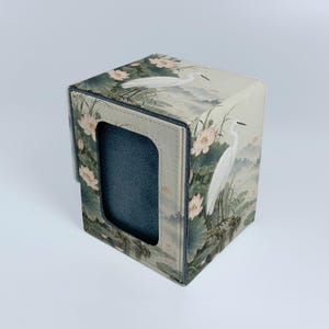 Può includere: Una scatola rettangolare con un motivo stampato di aironi bianchi e fiori di loto rosa su sfondo beige chiaro. Il pannello frontale presenta un'apertura rettangolare che rivela un rivestimento interno grigio scuro dalla superficie ruvida.