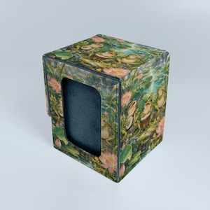 Könnte beinhalten: Eine quadratische Deck-Box mit einer farbenfrohen Illustration von Fröschen, die zwischen rosa Lotusblüten und grünen Seerosenblättern Musikinstrumente spielen. Die Box hat einen rechteckigen Ausschnitt, der ein dunkelgraues Innenfutter zeigt.