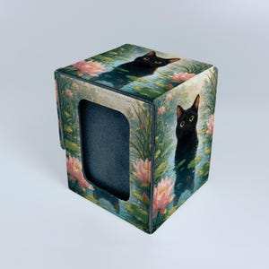 Op de afbeelding: Een vierkante opbergdoos met een print van een zwarte kat die uit een vijver vol roze lotusbloemen en groene waterlelies komt. De doos heeft een donkergrijze binnenvoering en een rechthoekige uitsparing aan de voorzijde van het paneel.