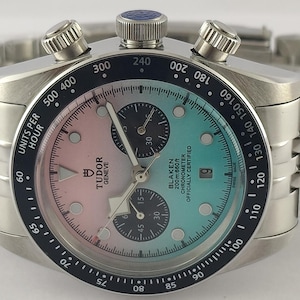 Peut inclure: Une montre chronographe Tudor en acier inoxydable avec un cadran dégradé rose et bleu sarcelle, des sous-cadrans noirs et une lunette tachymètre noire. La montre présente un bracelet à maillons argenté, un guichet de date et des index blancs.