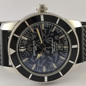 Può includere: Un orologio Breitling Superocean con cinturino in gomma nera testurizzata e cassa in acciaio inossidabile color argento. Il quadrante nero presenta un sottile motivo floreale, lancette luminose, indici argentati, una finestrella data e la scritta Breitling 1884.