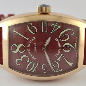 Op de afbeelding: Een tonvormig polshorloge van roségoud met een dieprode wijzerplaat met grote, niet-opeenvolgende witte Arabische cijfers. Het horloge heeft een rode horlogeband met alligatorstructuur, een gouden kroon en de tekst Franck Muller Geneve Crazy Hours Swiss Made.