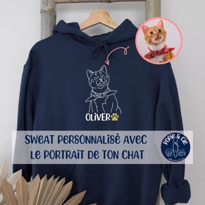 Puede incluir: Una sudadera con capucha azul marino que presenta una ilustración de línea blanca de un gato con un pañuelo. Debajo del gato, el nombre OLIVER está impreso en blanco, seguido de una huella dorada. Un recuadro circular muestra al gato real.