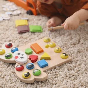 May include: A wooden puzzle board featuring a white face cutout and various geometric shapes in red, blue, yellow, green, and orange. A small wooden fishing rod with a string and magnet is used to lift the colorful pieces from the board surface.
