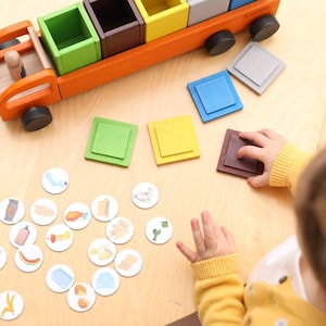 Könnte beinhalten: Ein hölzerner Spielzeuglaster mit orangefarbenem Rahmen und schwarzen Rädern, der vier offene Fächer in Grün, Braun, Gelb und Grau aufweist. Neben dem Laster liegen passende quadratische Holzdeckel und eine Sammlung kleiner weißer runder Spielmarken mit Aufdrucken.