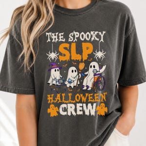 May include: A charcoal gray t-shirt featuring the text "THE SPOOKY SLP HALLOWEEN CREW" in orange and white. The graphic depicts three white ghosts: one with a purple hat and walker, one holding a broom, and one in a wheelchair.