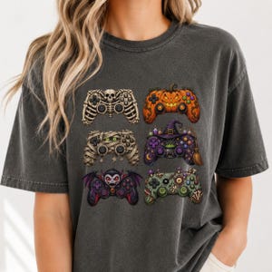 Op de afbeelding: Een antracietgrijs T-shirt met korte mouwen en een grafische print van zes videogamecontrollers met Halloween-thema's, waaronder een skelet, een pompoen, een mummie, een heks, een vampier en een op zombies geïnspireerde esthetiek.