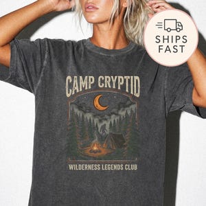 Può includere: Una t-shirt a maniche corte grigio antracite con una stampa grafica raffigurante una scena boschiva con un falò, una tenda, una luna crescente e la sagoma di una creatura. Il testo recita CAMP CRYPTID e WILDERNESS LEGENDS CLUB in stile vintage.