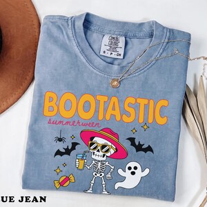 May include: A blue jean colored t-shirt featuring the text BOOTTASTIC and summerween in orange and pink. The graphic includes a skeleton wearing a pink hat and sunglasses holding a drink, a white ghost, a spider, bats, stars, and candy.
