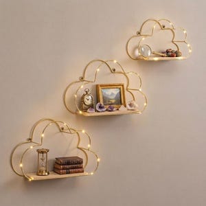 May include: Three cloud-shaped wall shelves made of gold-toned metal wire with integrated warm LED string lights. Each shelf features a flat wooden base. The shelves hold decorative items including a small clock, a framed picture, books, and stones.