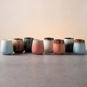 Op de afbeelding: Een verzameling van acht keramische bekers op een rij, met diverse afwerkingen waaronder effen matte pasteltinten, gespikkelde texturen en kleurverlopen in de tinten blauw, bruin, groen, roze, wit en zwart.
