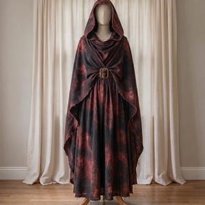 Könnte beinhalten: Ein bodenlanger Kapuzenumhang mit einem dunkel schwarzen und tiefroten, fleckigen Batikmuster. Das Kleidungsstück verfügt über eine große, auffällige rechteckige Schnalle in Bronzefarbe an der Brust, die den Stoff drapiert hält.