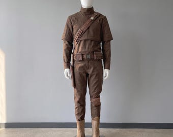Peut inclure: Une chemise marron à manches longues avec un col montant, un pantalon assorti et une ceinture utilitaire marron avec des poches. Une bandoulière marron avec de petites boucles cylindriques traverse la poitrine. L'ensemble inclut un étui et des bottes marron clair.