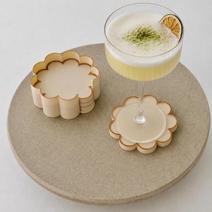 May include: A set of light-coloured, flower-shaped coasters with scalloped edges, stacked together next to a single coaster supporting a ribbed glass filled with a pale yellow cocktail, topped with foam, green powder, and a thin slice of dried lime.