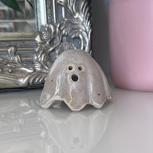May include: A small, beige, ceramic ghost figurine with a glossy, speckled finish. The ghost features two small circular eyes and a larger circular mouth opening, creating a hollow, expressive face. The base has a scalloped, draped edge.