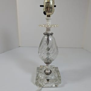 May include: A table lamp base featuring a clear glass body with a spiral twist design, a square glass base, and white accents. The top includes a metallic lamp socket with visible text reading SUN-LITE 660W 250V MADE IN CHINA.