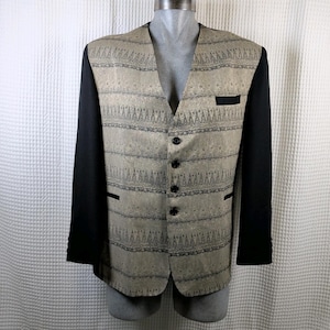 May include: A blazer with solid black sleeves and a tan front panel featuring a repeating dark grey geometric pattern. The garment has a V-neckline, four black buttons down the centre, a single chest pocket, and two lower pockets with black trim.
