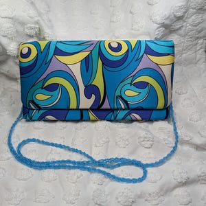 Può includere: Una pochette rettangolare caratterizzata da un vivace motivo astratto di forme vorticose nei toni del blu, verde lime, viola e crema. La borsa include una lunga e sottile tracolla di perline azzurre fissata ai lati della struttura.
