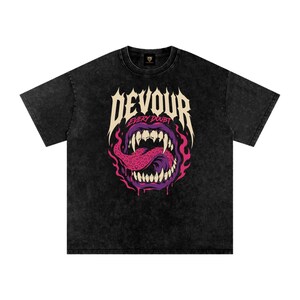 以下が含まれることがあります： 黒いアシッドウォッシュ加工のTシャツ。鋭い歯と斑点のあるピンクの舌を持つ紫色の口のグラフィックが描かれています。口の上には大きなクリーム色のフォントで「DEVOUR」と書かれ、その下に小さなピンクの文字で「EVERY DOUBT」と記されています。