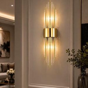 May include: A vertical wall sconce featuring a central gold metal band that holds multiple clear glass rods extending upward and downward. The fixture emits a warm, glowing light from within the glass rods, creating a bright, illuminated effect.