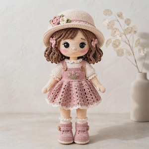 Könnte beinhalten: Eine handgefertigte Häkelpuppe mit braunem Haar, die einen cremefarbenen Hut mit rosa und weißen Blumen, ein weißes Kurzarmshirt, ein altrosa Trägerkleid mit Blumenstickerei sowie passende rosa Stiefel mit kleinen Knöpfen trägt.