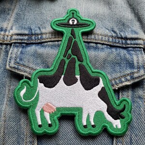 May include: An embroidered patch featuring a black and white cow with a pink udder, standing in a grazing position. A green, elongated shape extends upward from the cow's back, topped with a small black and white alien spaceship graphic.