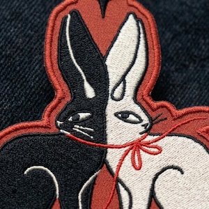 May include: An embroidered patch featuring two rabbits, one black and one white, facing each other. The rabbits are connected by a thin red thread tied in a bow around their necks. The entire design is set against a dark red background.