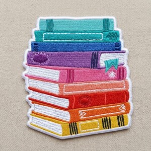 May include: An embroidered patch featuring a stack of eight books in various colors including teal, blue, purple, pink, orange, red, and yellow. Each book spine shows distinct textures and patterns, with white thread detailing the pages.