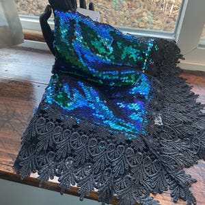 May include: A decorative scarf with iridescent blue and green sequins. The scarf is trimmed with a black lace border. The scarf is displayed on a dark wooden surface near a window.