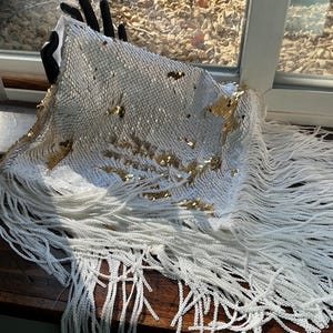 May include: White sequin fabric with gold accents and long white fringe. The fabric is draped over a black hand. The sequins are small and closely spaced, creating a shimmering effect. The fringe hangs down, adding texture and movement.